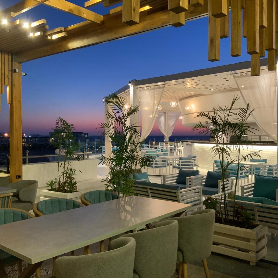 Sunrise Alex Avenue Hotel Alexandria Exterior photo The photo shows a stylish outdoor dining area or terrace set against a beautiful sunset. There are several tables and chairs arranged neatly under a covered structure, with a backdrop of soft white curtains. The space is decorated with potted plants,