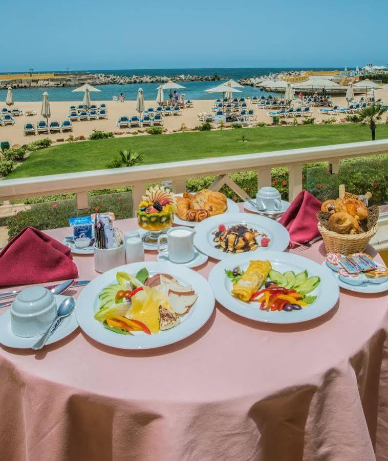 Sunrise Alex Avenue Hotel Alexandria Exterior photo The photo shows a beautifully set breakfast table on a balcony with a scenic view of a beach. There are several plates of food arranged attractively. One plate features an assortment of fruits, including slices of melon, berries, and other colorful f