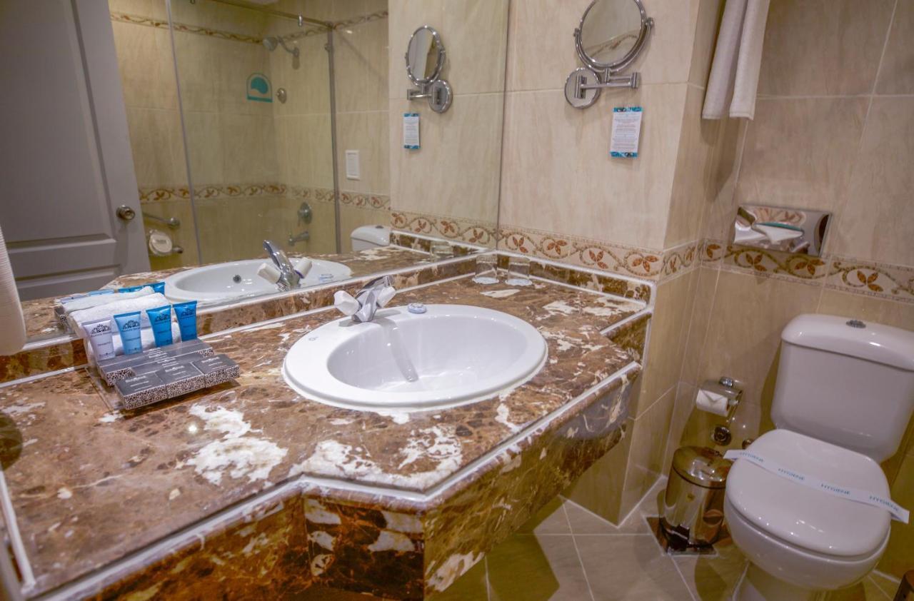 Sunrise Alex Avenue Hotel Alexandria Exterior photo The photo shows a bathroom with a marble countertop featuring a round sink. There is a large mirror behind the sink, reflecting the space. The backsplash has decorative tile accents, and on the counter, there are several toiletries presented in blue 