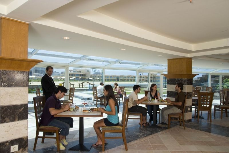 Sunrise Alex Avenue Hotel Alexandria Restaurant photo The photo shows an indoor dining area, possibly in a restaurant or café. There are several tables with people sitting at them. Two groups of diners are engaged in conversation while enjoying their meals. A server is also present, attending to one of 