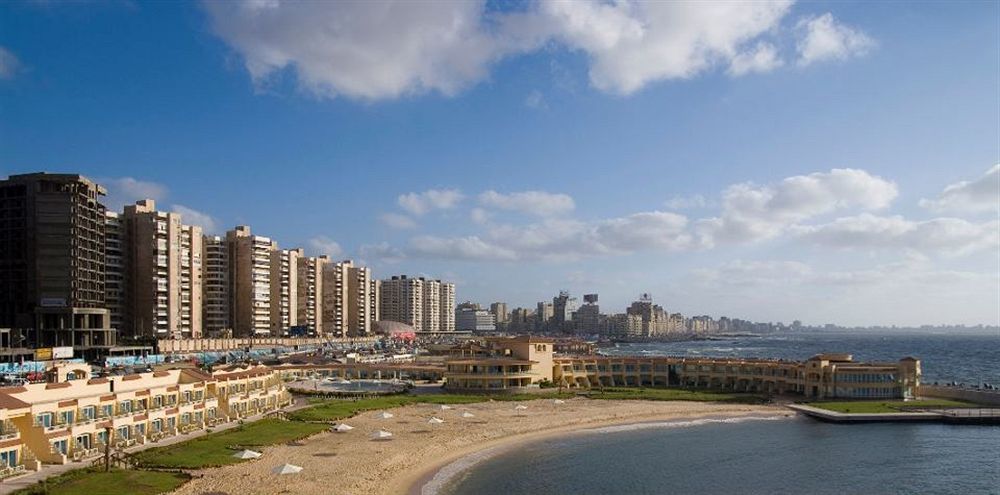 Sunrise Alex Avenue Hotel Alexandria Exterior photo The photo shows a scenic coastal view featuring a sandy beach and a curved shoreline. In the foreground, there are several sun loungers and a landscaped area. Beyond the beach, there are multiple high-rise buildings lining the waterfront, indicating 