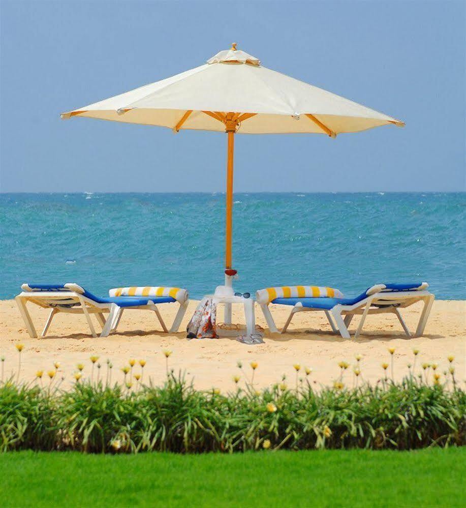 Sunrise Alex Avenue Hotel Alexandria Exterior photo The photo shows a serene beach scene featuring four lounge chairs arranged under a large umbrella. The chairs have blue and yellow stripes, and a small table is positioned between them. In the background, the turquoise ocean stretches out under a cle