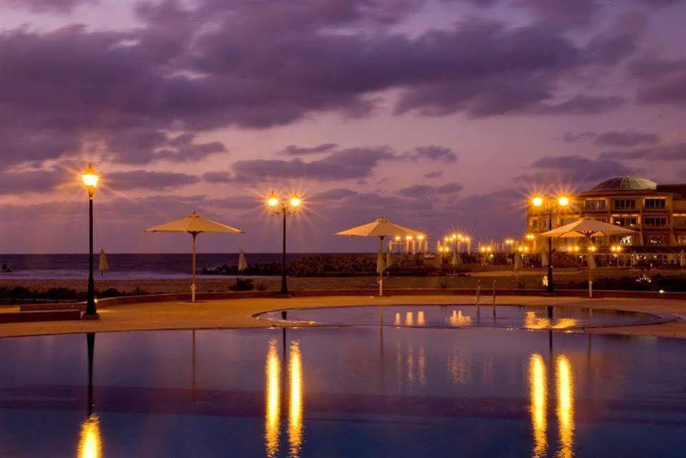 Sunrise Alex Avenue Hotel Alexandria Exterior photo The photo depicts a serene evening scene at a coastal location. In the foreground, there is a calm pool of water that reflects the surrounding environment. To the sides, there are several umbrellas and lamp posts that emit a warm glow, enhancing the 