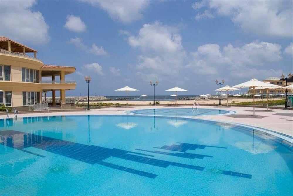 Sunrise Alex Avenue Hotel Alexandria Exterior photo The image shows a beautiful outdoor scene featuring a large swimming pool with clear blue water, surrounded by a well-maintained area. In the background, there are sun umbrellas set up, indicating a relaxation area, possibly by the beach or a resort.