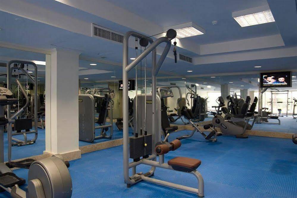 Sunrise Alex Avenue Hotel Alexandria Exterior photo The photo shows a modern gym interior. There is a cable machine in the foreground, which is used for resistance training, featuring two adjustable seats. The flooring is covered with blue mats, and in the background, various other gym equipment such 