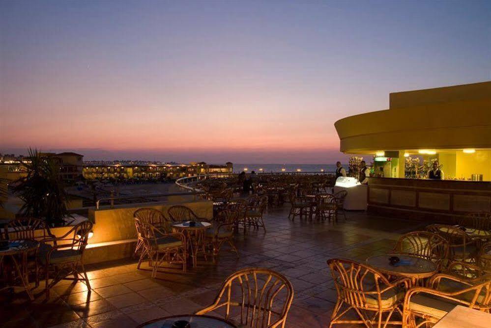 Sunrise Alex Avenue Hotel Alexandria Exterior photo The photo depicts a picturesque outdoor setting during twilight. In the foreground, there are several wicker chairs and tables arranged around a spacious area. The floor is made of tiles, and the lighting creates a warm ambiance. In the background, a