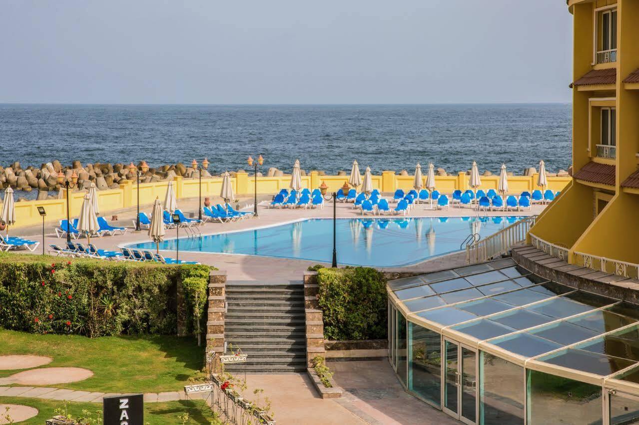 Sunrise Alex Avenue Hotel Alexandria Exterior photo The photo depicts a scenic view of a pool area adjacent to the ocean. In the foreground, there is a large swimming pool surrounded by numerous blue lounge chairs and umbrellas, indicating a relaxing setting. There are steps leading down from a grassy