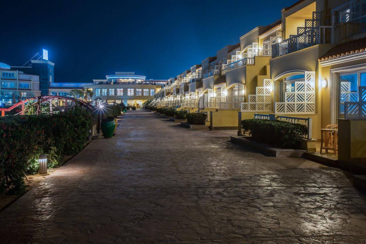 Sunrise Alex Avenue Hotel Alexandria Exterior photo The photo depicts a nighttime scene of a resort or hotel area. There is a wide, well-paved pathway lined with well-lit structures, likely guest rooms or villas, on either side. The buildings have a warm yellow hue, and the pathway is illuminated by s