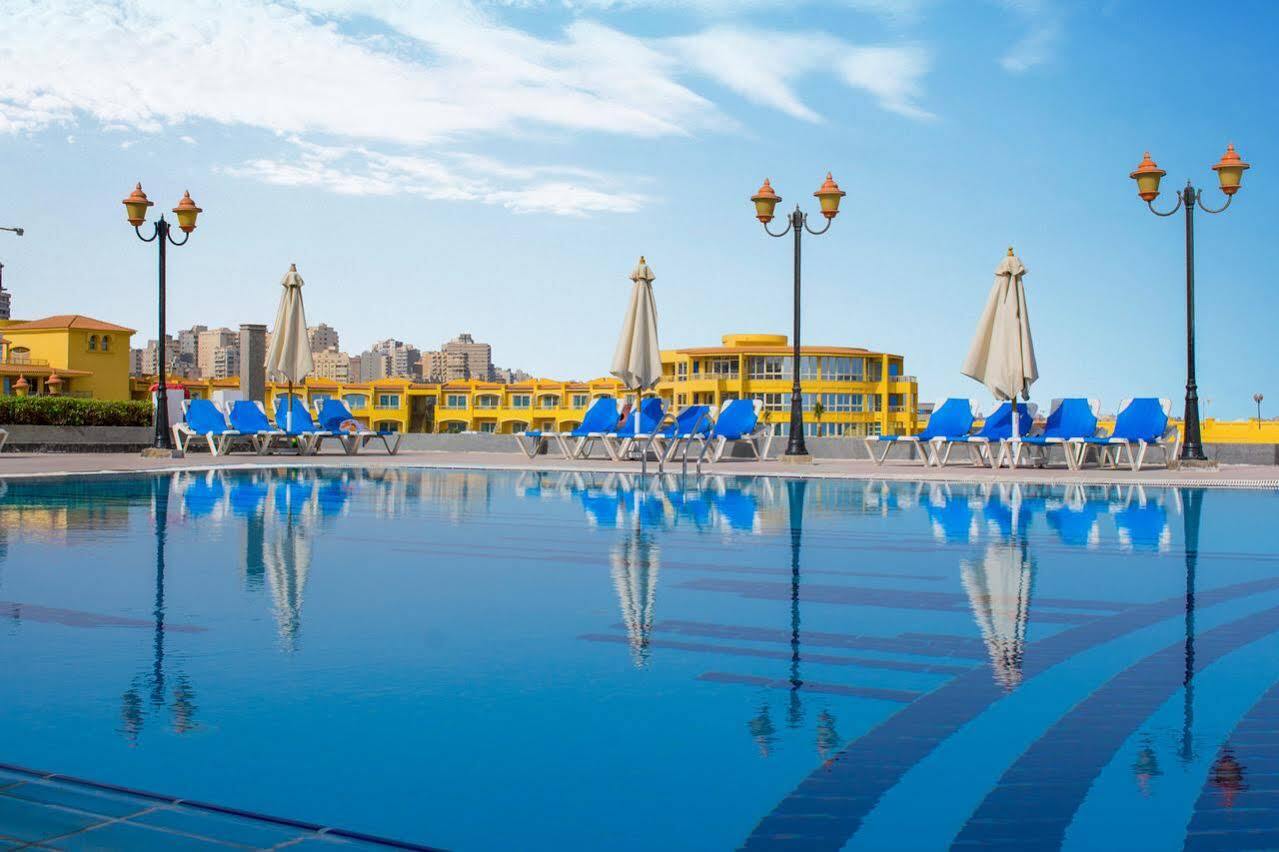 Sunrise Alex Avenue Hotel Alexandria Exterior photo The photo shows a swimming pool with clear blue water reflecting the sky. Surrounding the pool are several blue lounge chairs and large umbrellas. In the background, there's a yellow building that appears to be a hotel or resort, and lampposts that a