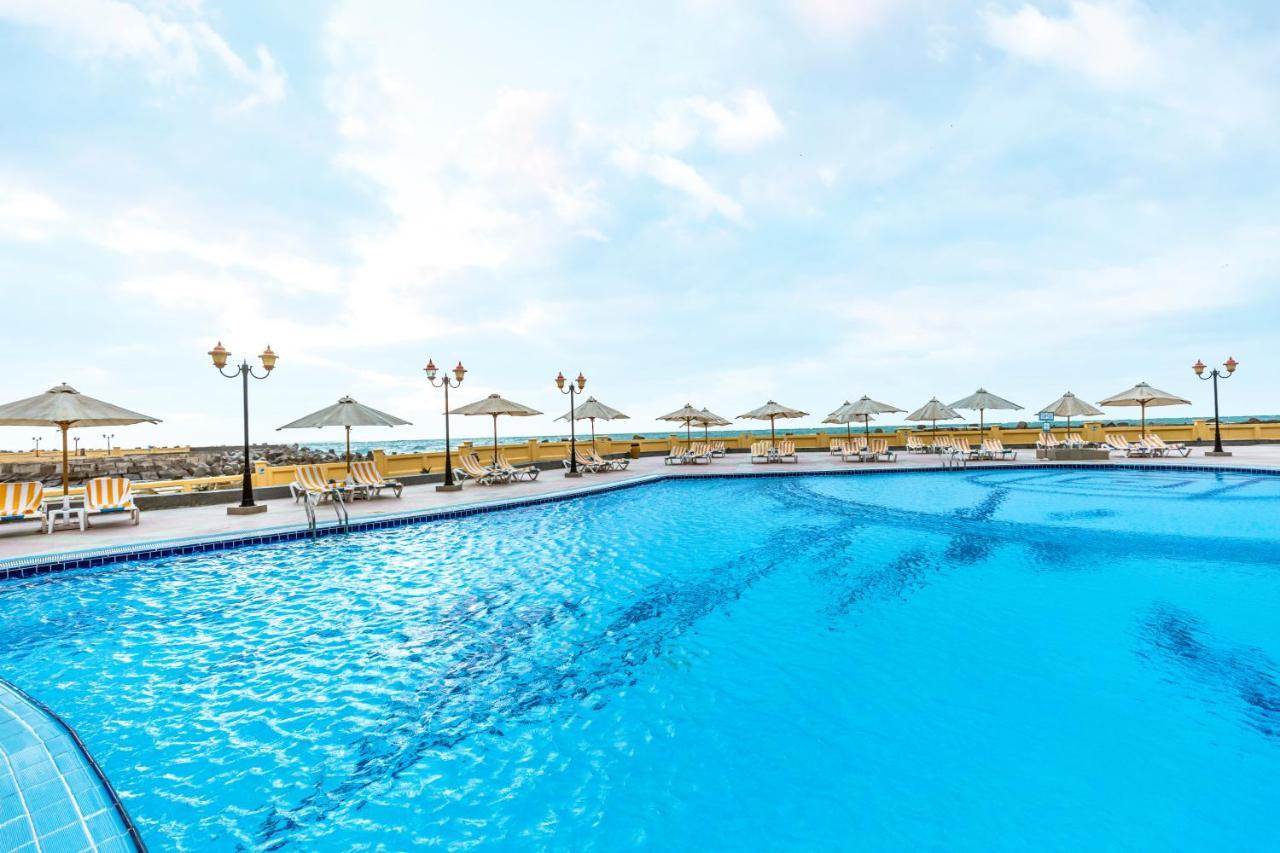 Sunrise Alex Avenue Hotel Alexandria Exterior photo The photo shows a large swimming pool with crystal clear blue water. Surrounding the pool are several sun loungers and umbrellas, providing shade for relaxation. In the background, there appear to be more lounge chairs and additional umbrellas, sugge